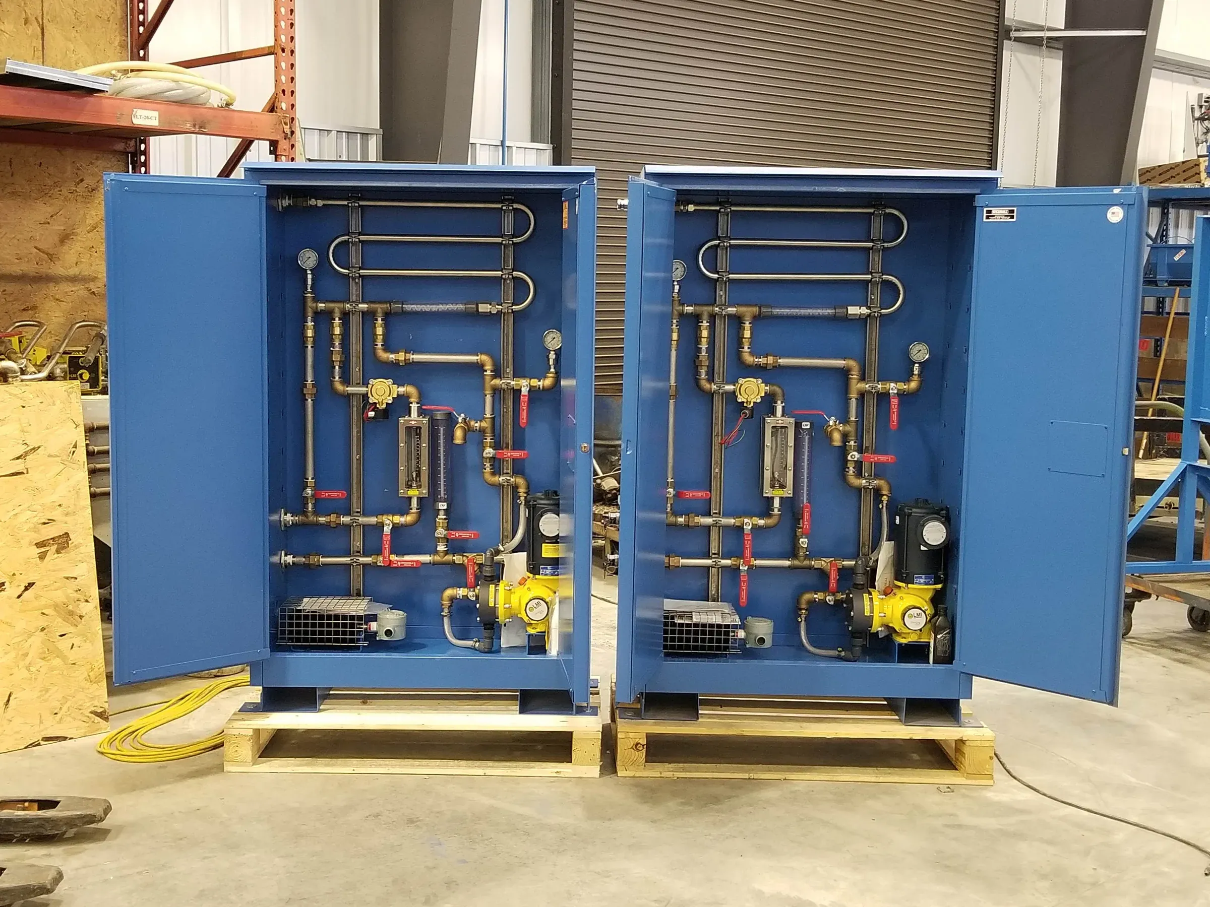 Two blue chemical feed cabinets with their doors open on a shop floor, showing gauges, rotameters, ball valves and metering pumps