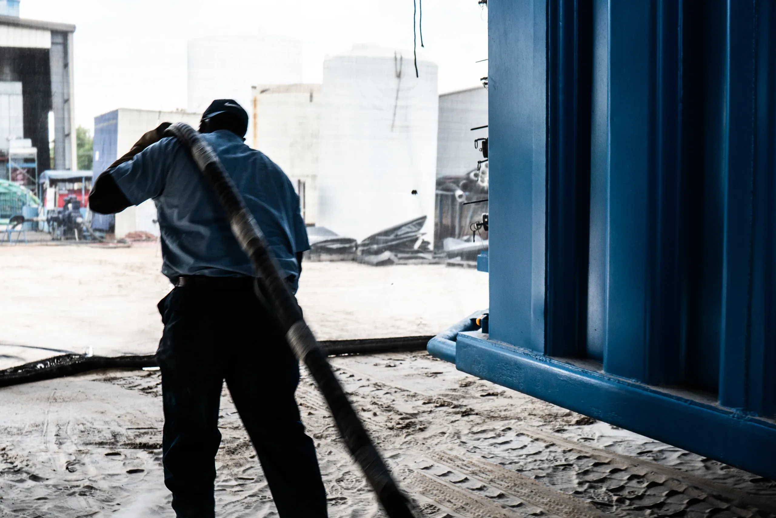 Technician hauling a suction hose over his shoulder across a sand pad beside a blue steel treatment tank