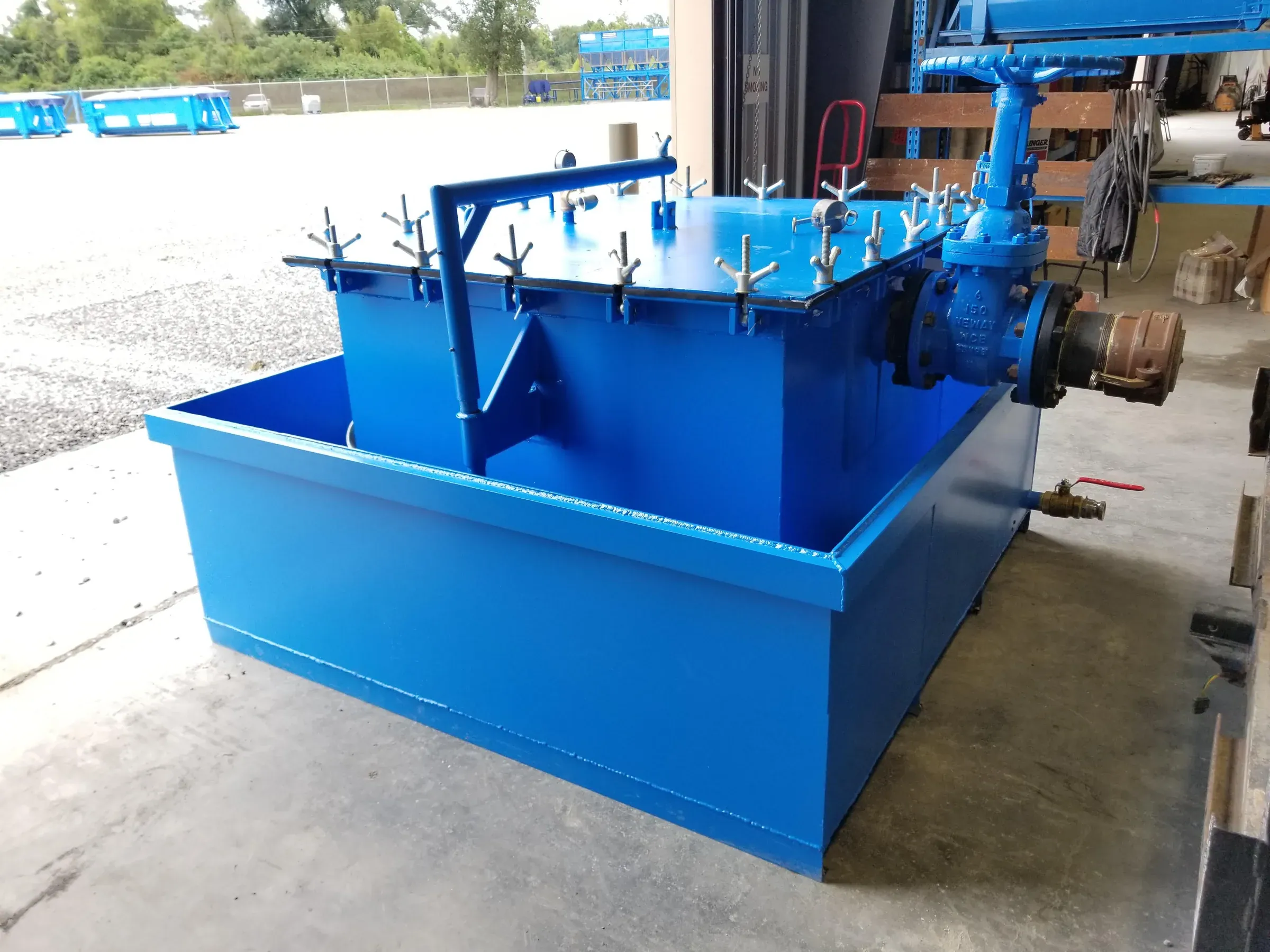 Custom-fabricated blue filter box with bolted access lid, overflow weir, gate valve and cam-lock inlet staged in the shop doorway