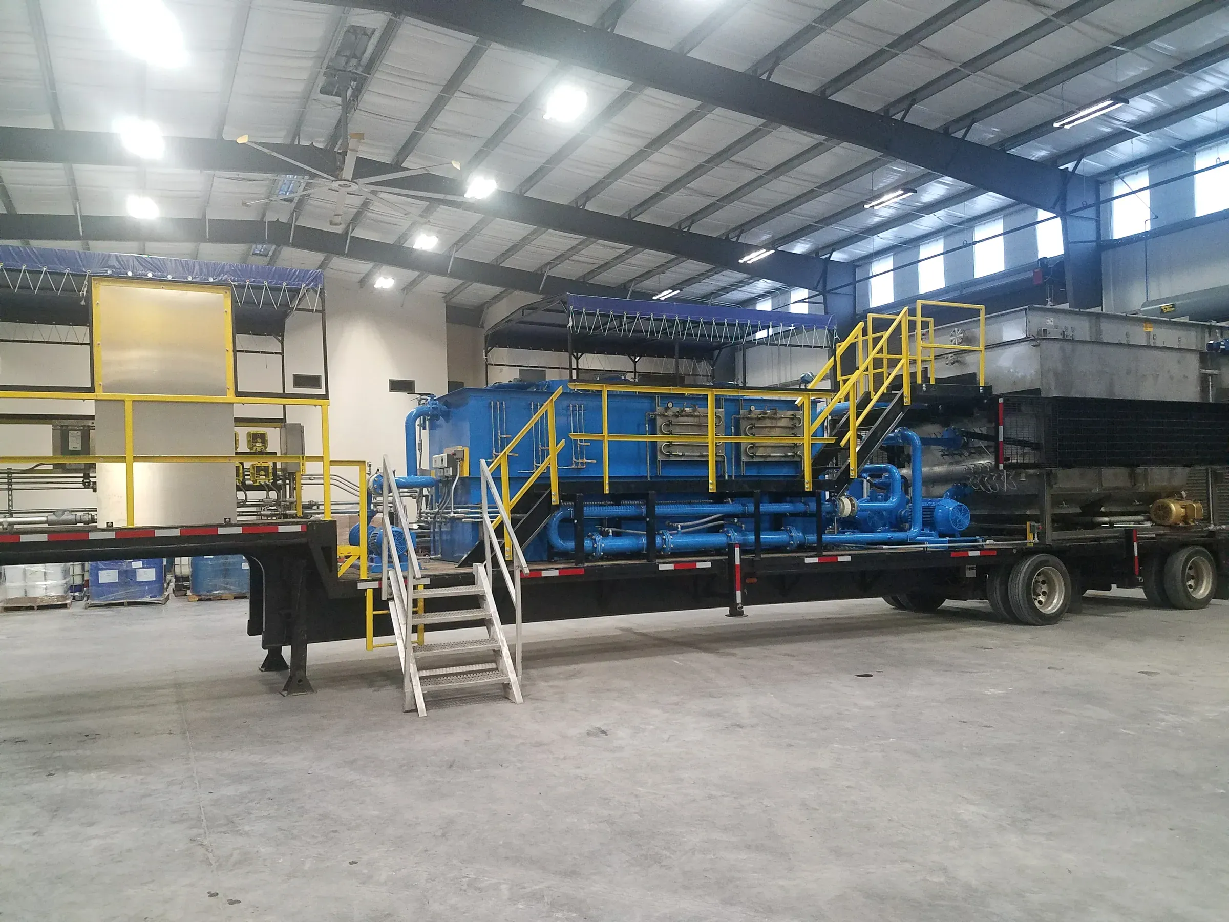 Trailer-mounted blue dissolved air flotation and clarifier treatment train with yellow access stairs and catwalk, parked inside a bright fabrication bay