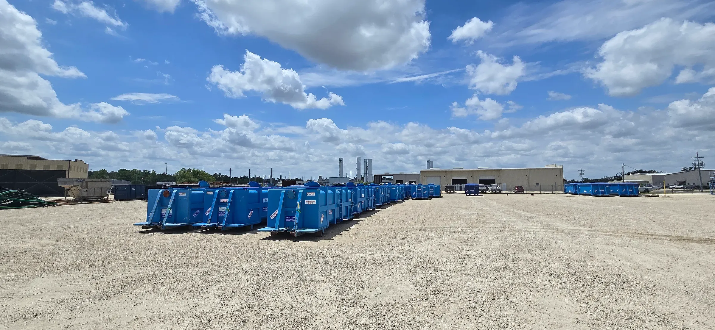 Line of blue roll-off containers staged across the gravel yard under a wide cumulus sky
