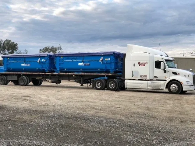 Two tarped blue dewatering boxes riding a flatbed trailer behind a white tractor, staged on a gravel yard