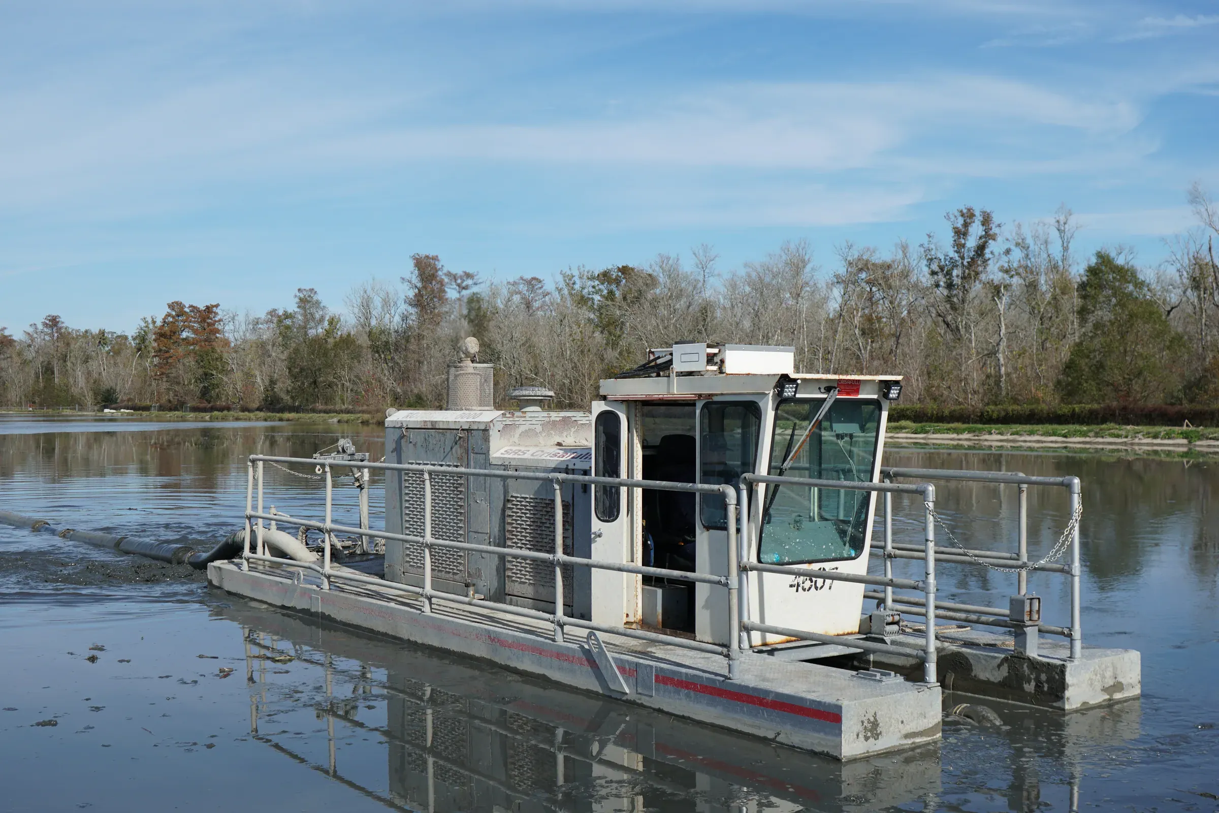 Pontoon work boat with an enclosed cab moving across the flat surface of an industrial lagoon, bare treeline on the far bank