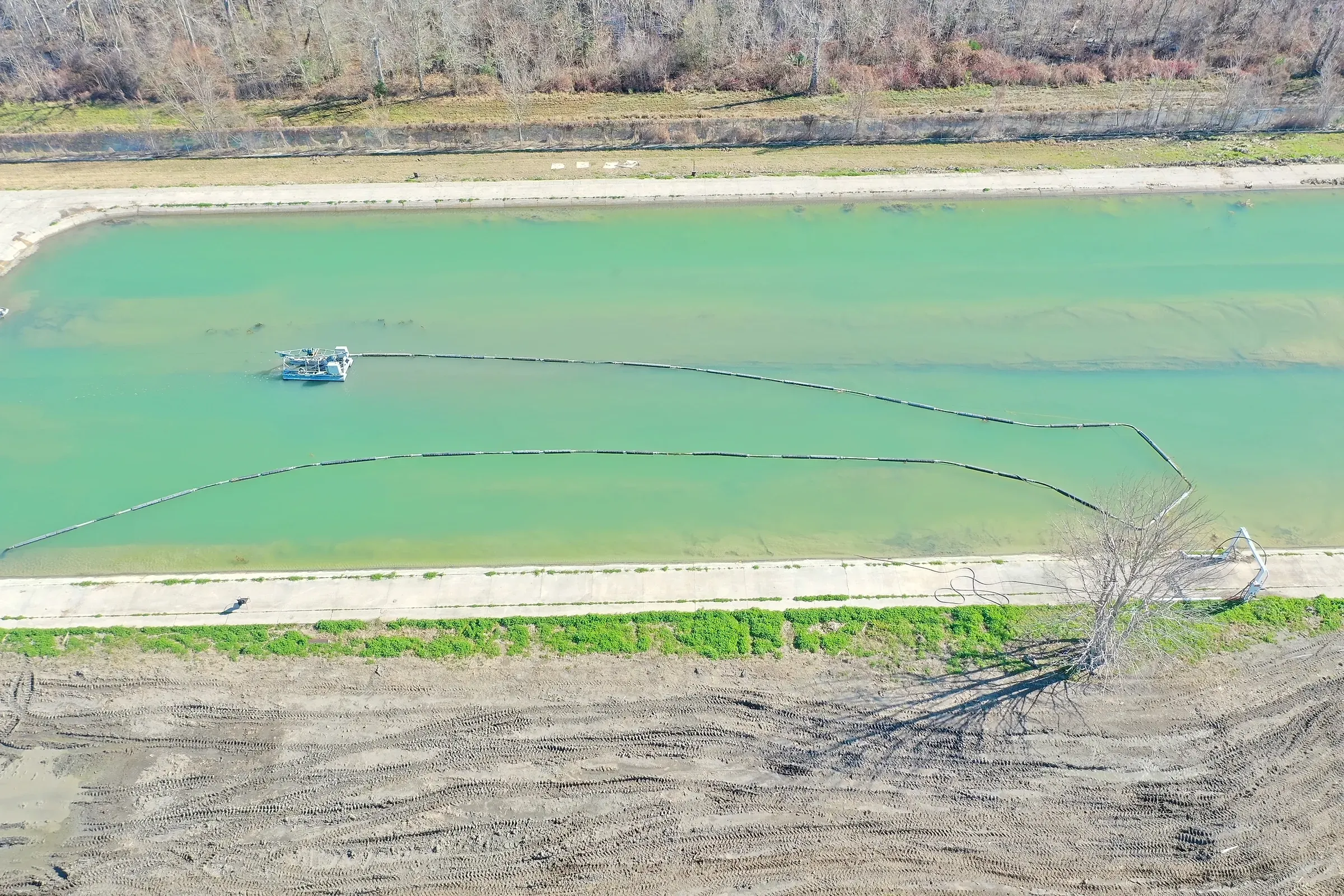 Aerial view of a cutter-suction dredge working a jade-green industrial basin, its floating discharge line curving back to the bank