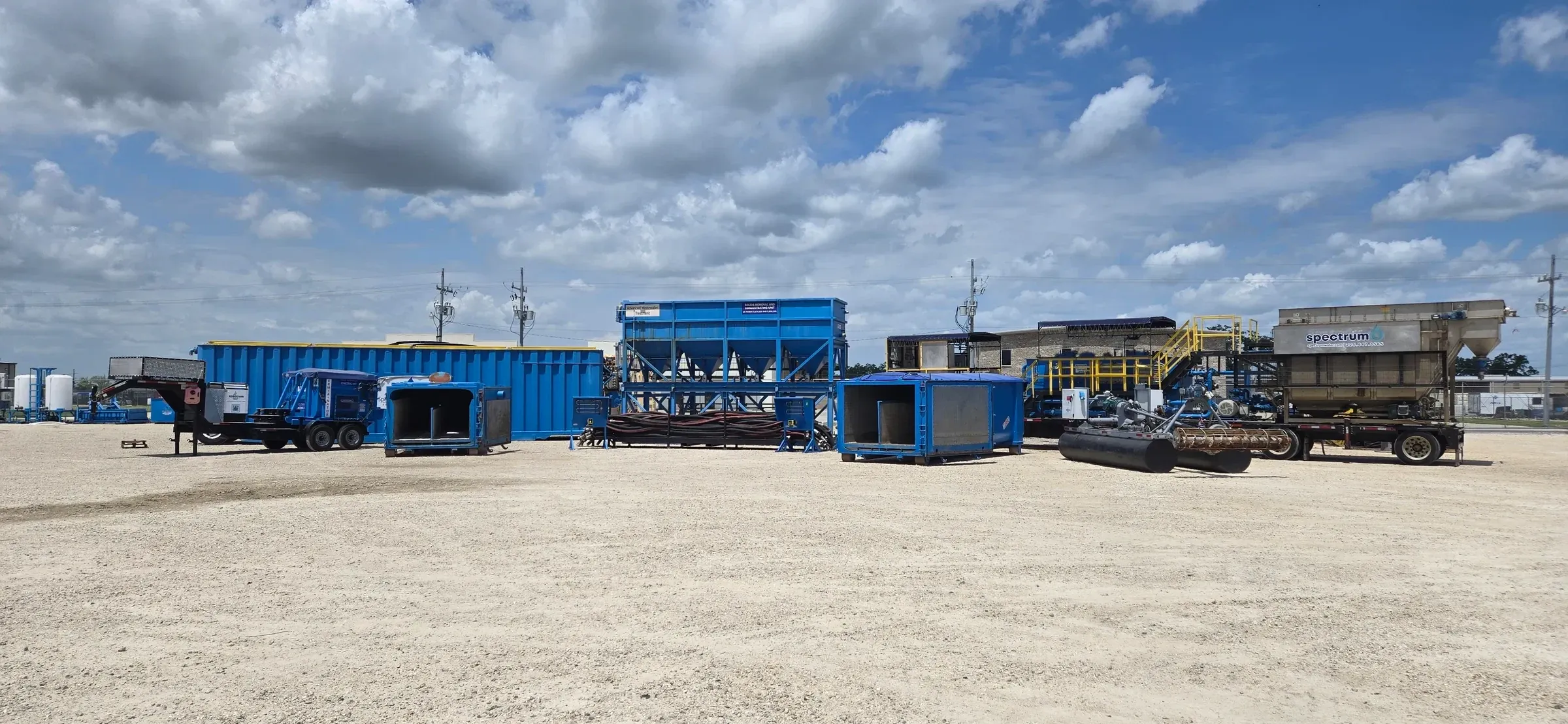 Wide view of the equipment yard with roll-off boxes, a solids concentrating unit and trailer-mounted treatment skids lined up under a big cloud-filled sky