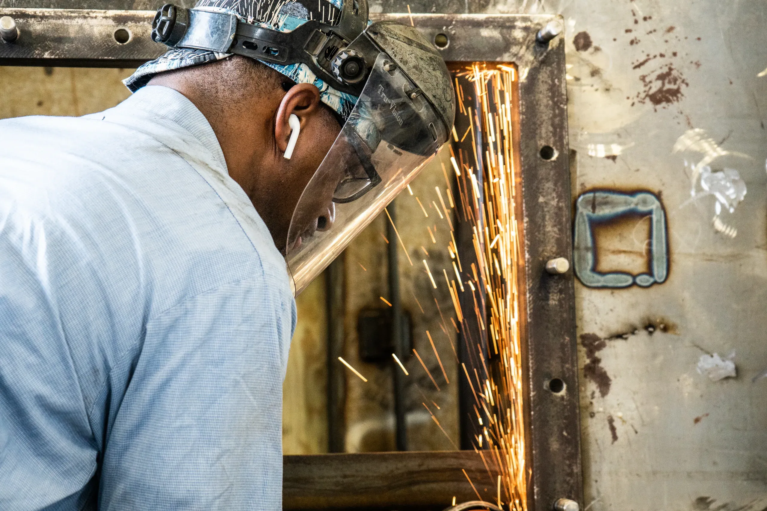 Close profile of a fabricator behind a clear face shield as an arc of grinder sparks sheets off the edge of the steel box he is working