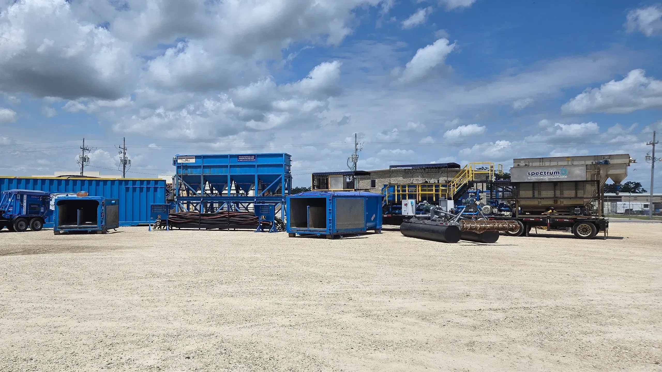 Spectrum equipment yard with roll-off boxes, a solids concentrating unit and trailer-mounted treatment skids lined up under a big cloud-filled sky