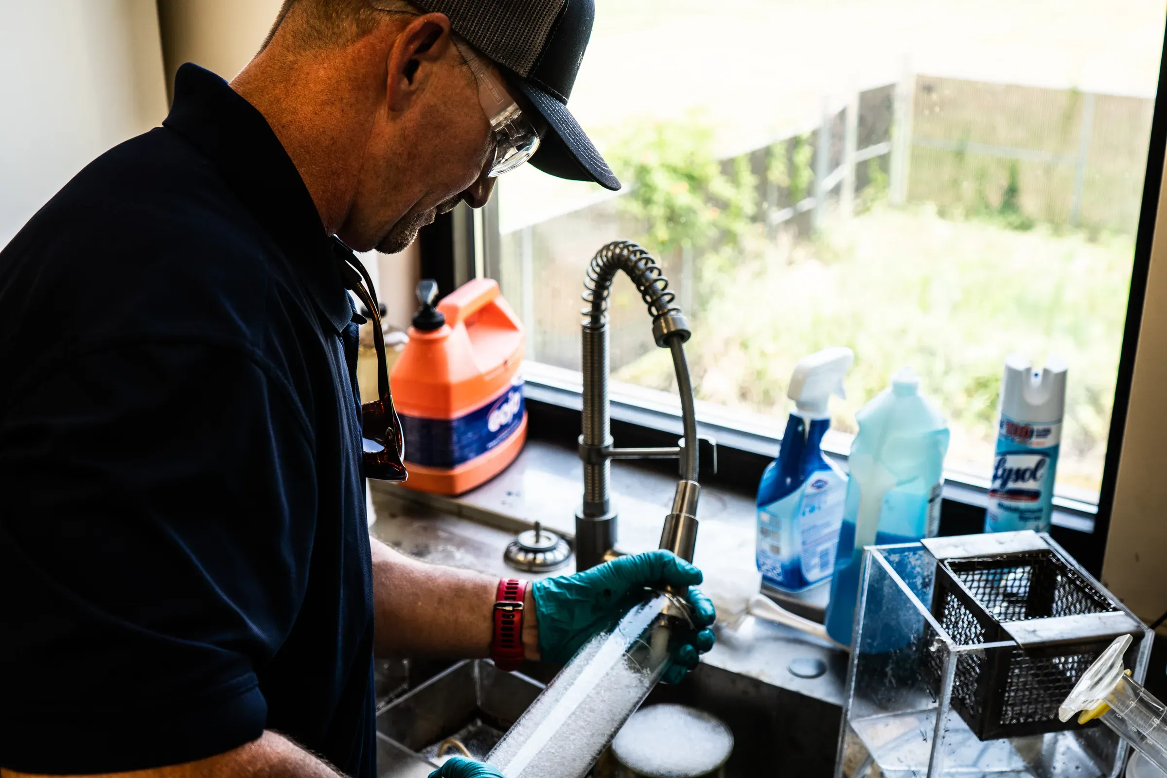 Technician in gloves rinsing a filter element at a lab sink beside a window, with sample glassware on the counter
