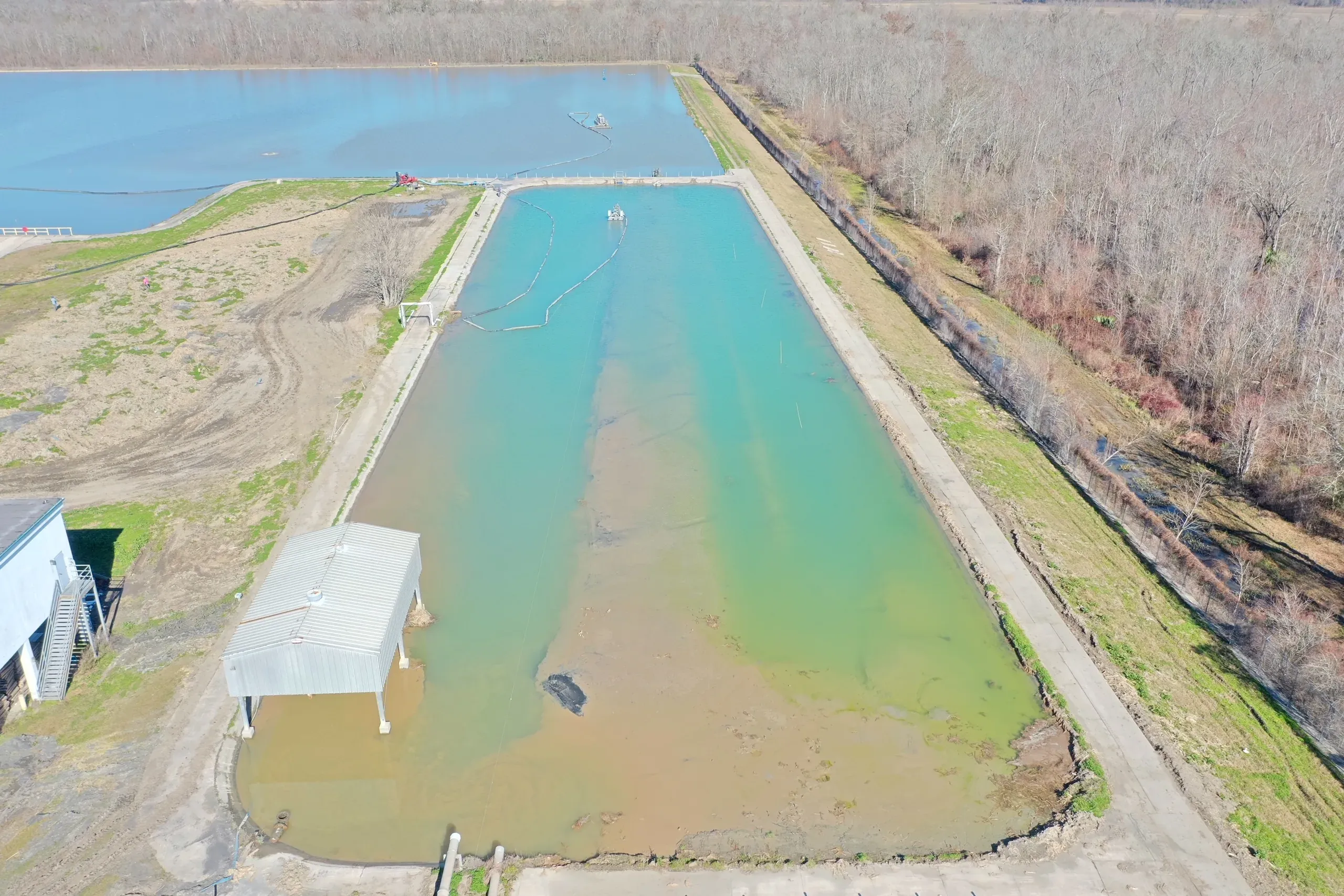 Aerial view of a lined industrial holding basin being dredged, teal clear water meeting ochre sediment, with a floating dredge and containment boom at the head of the cell