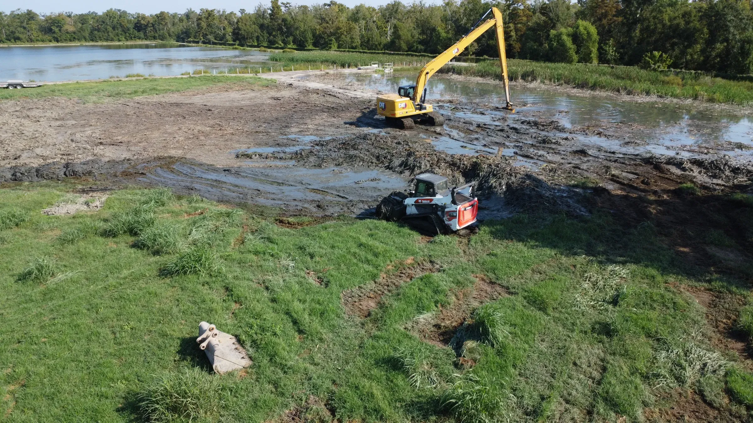 Aerial view of an excavator and a tracked loader clearing sediment from a drained lagoon beside an open lake