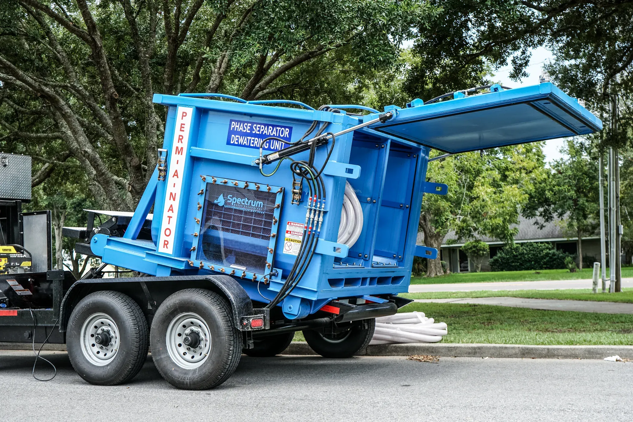 Trailer-mounted Spectrum Phase Separator dewatering unit with its lid raised and drain hose coiled beneath, parked under live oaks