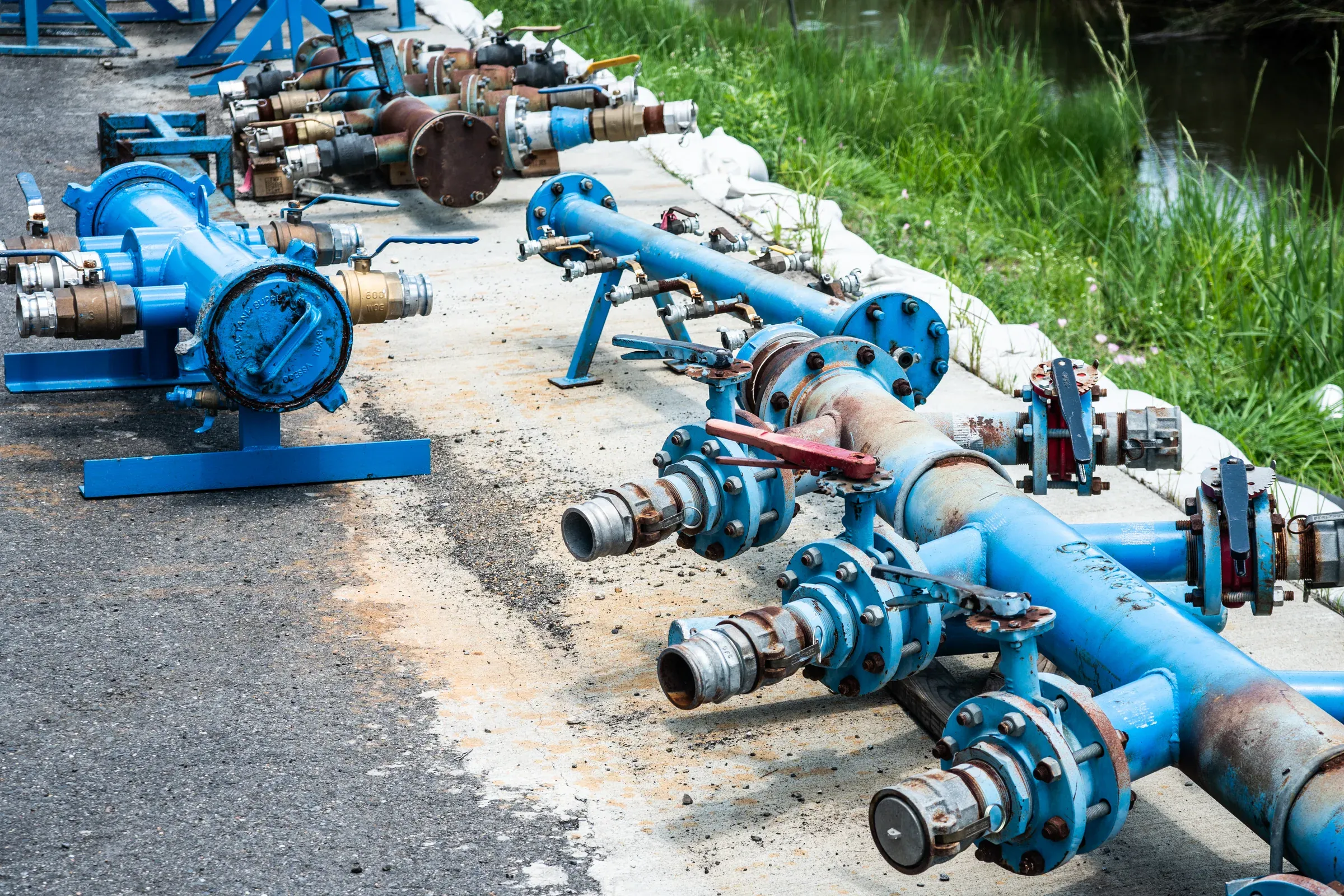 Rows of blue flanged pipe spools, ball valves and cam-lock fittings staged on a concrete pad beside a grassy canal bank