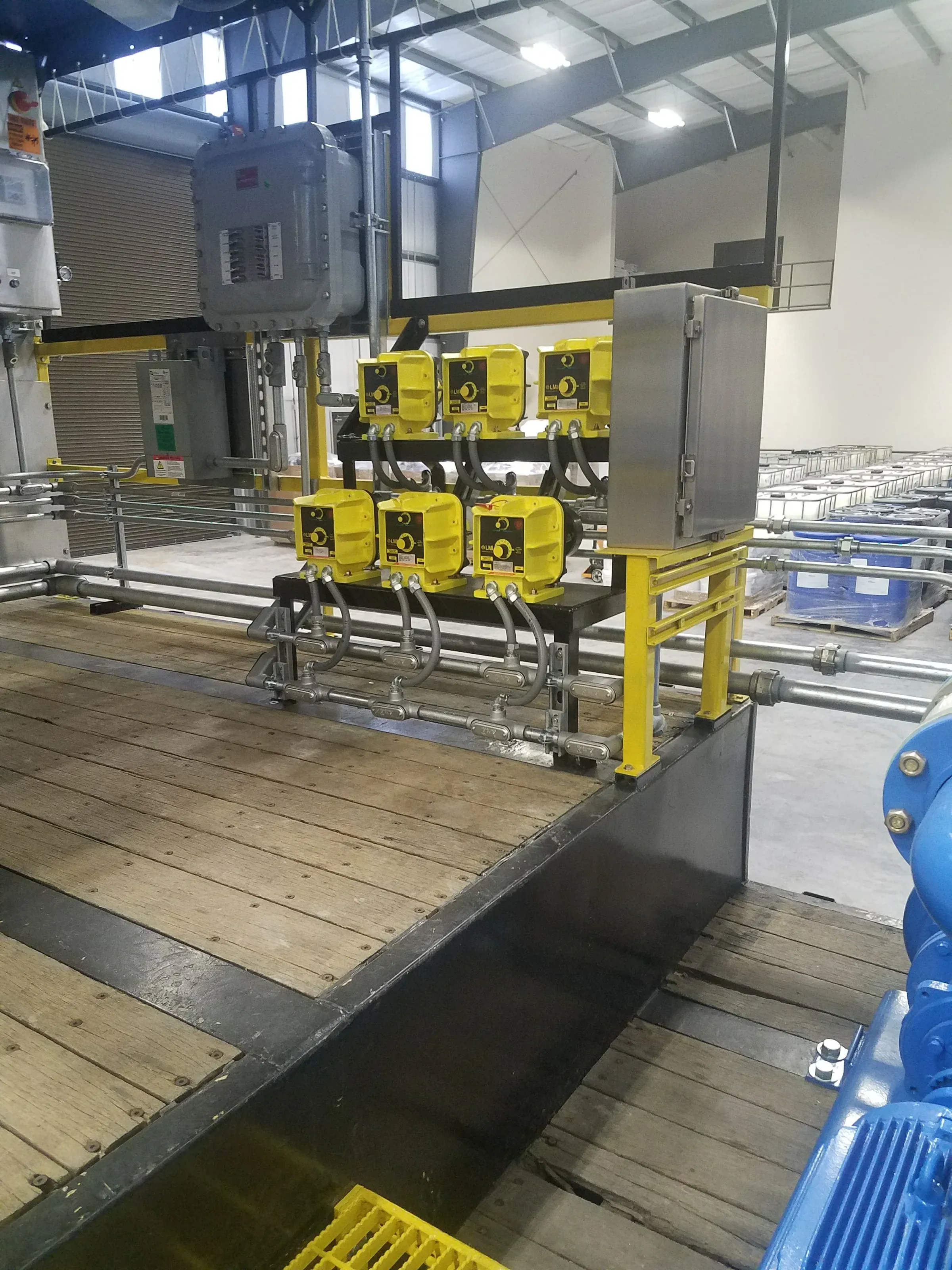 Bank of six yellow metering pumps mounted on a black skid above a treatment basin walkway inside a plant building