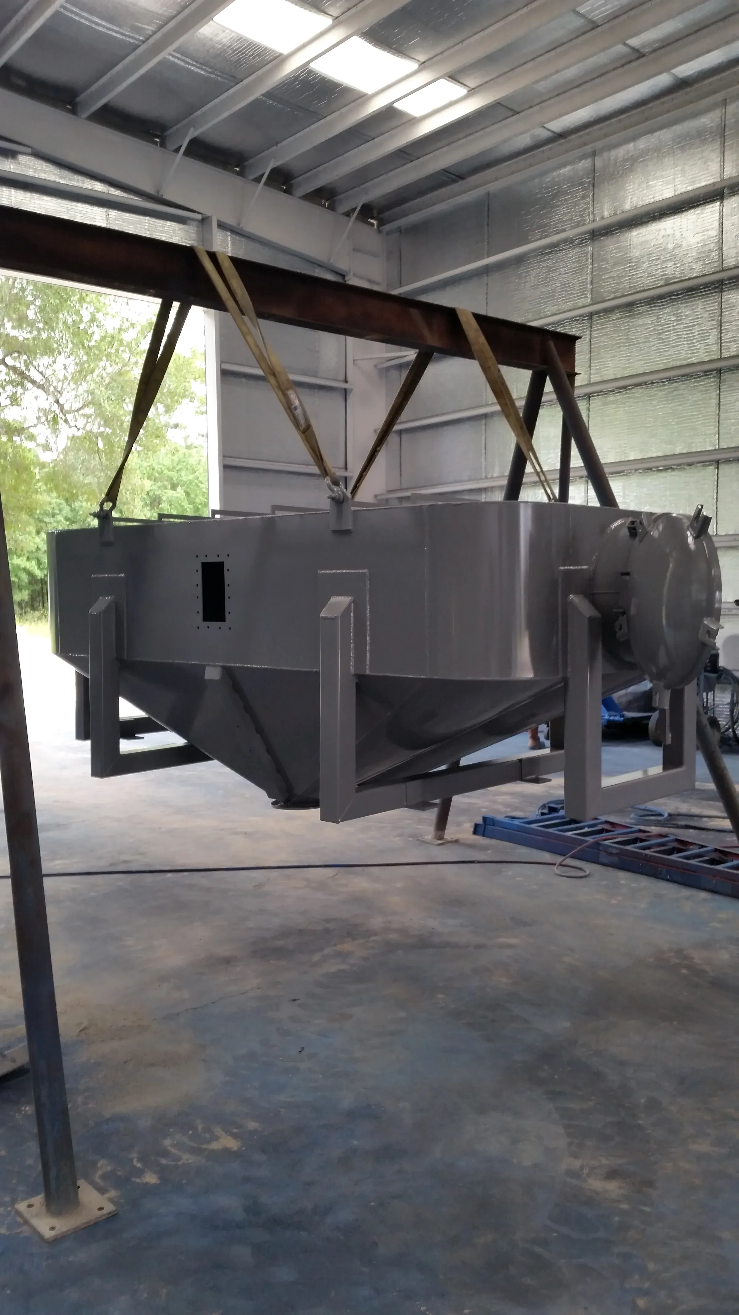 Freshly coated cone-bottom separator vessel slung from nylon straps on a lifting beam in the shop, hinged access hatch on the near end