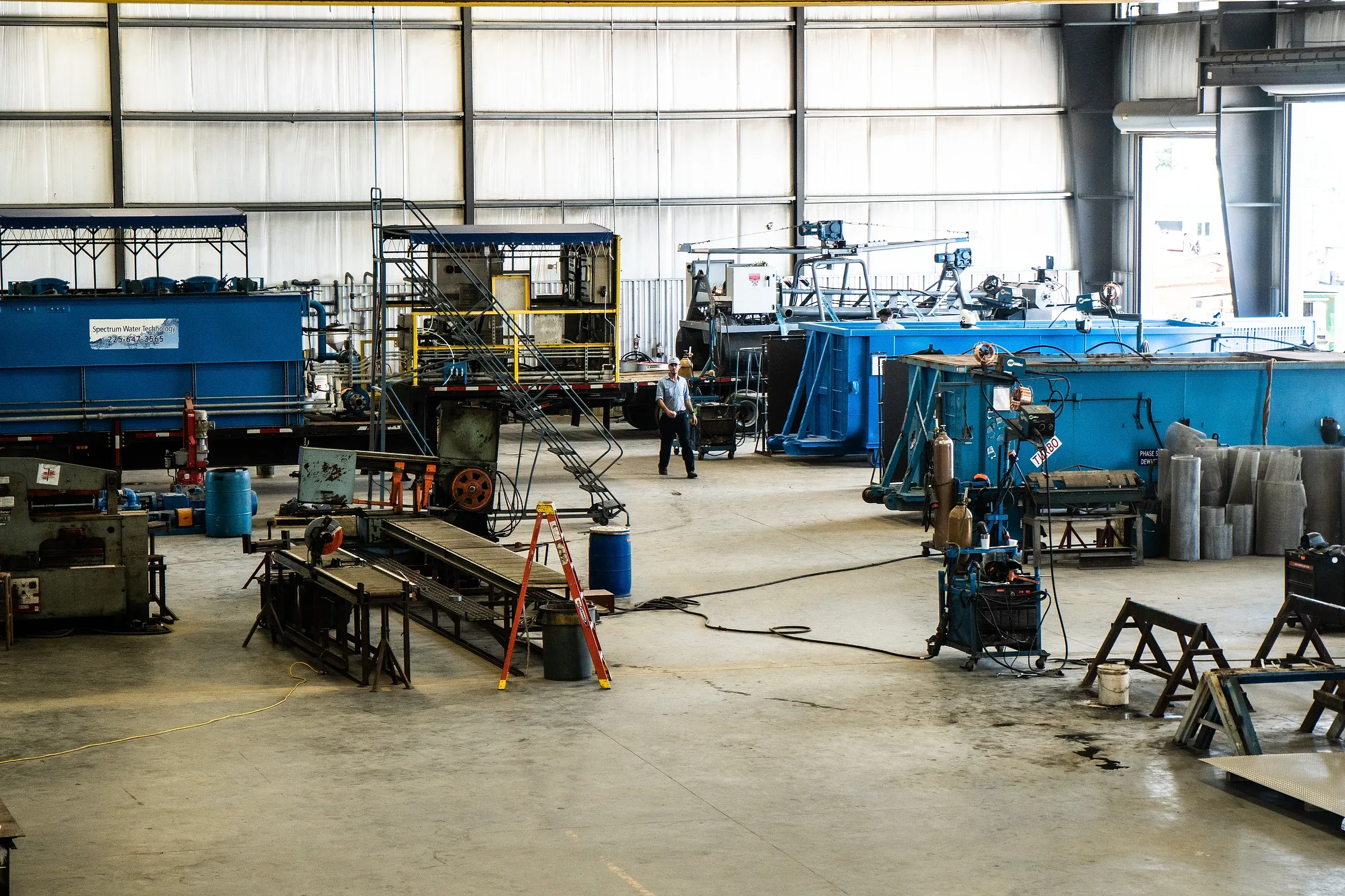 Open fabrication bay with blue treatment units, welding stations and a lone technician crossing the shop floor