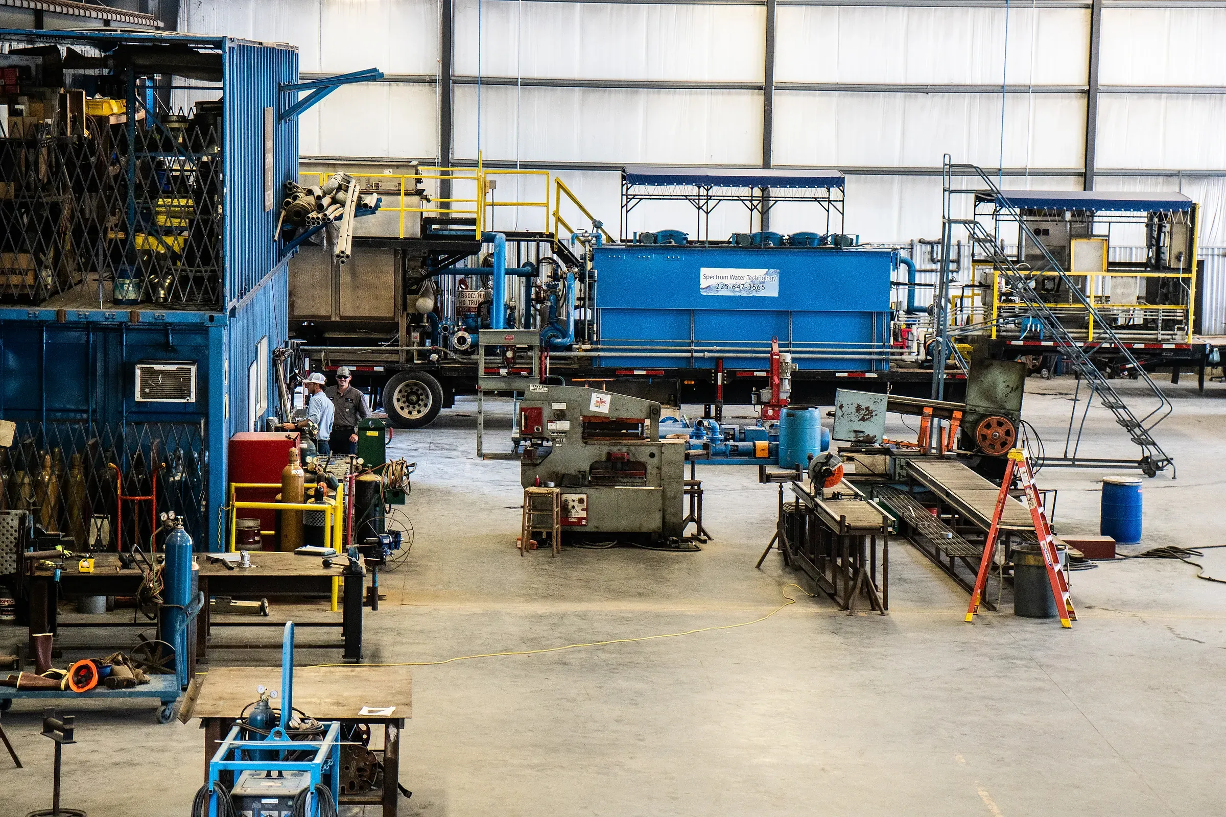 Wide view of the fabrication bay with trailer-mounted blue treatment units, ironworker press and welding stations