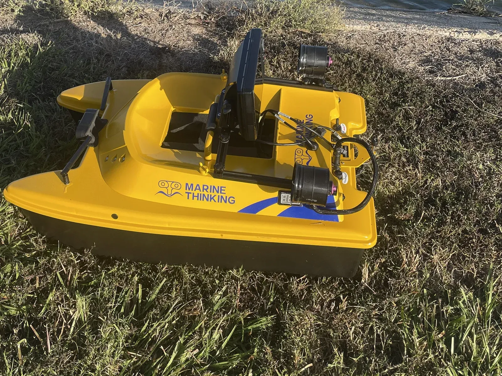 Close-up of the yellow remote survey boat on the grassy bank at the water's edge, sonar display and sensor pods mounted on deck