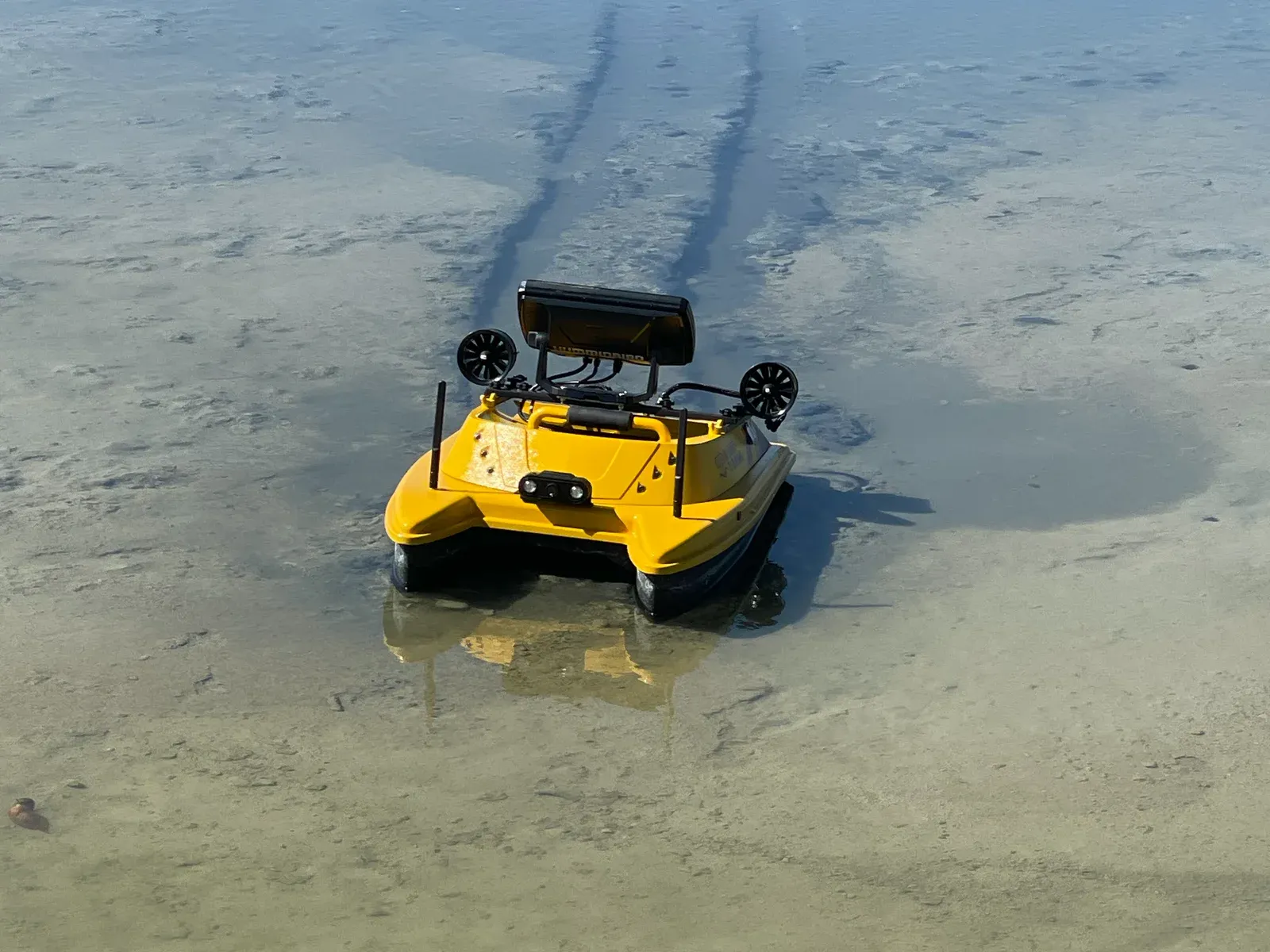 Yellow remote-controlled survey boat with a sonar head unit crossing a shallow pond, its track marks trailing through the sediment behind it