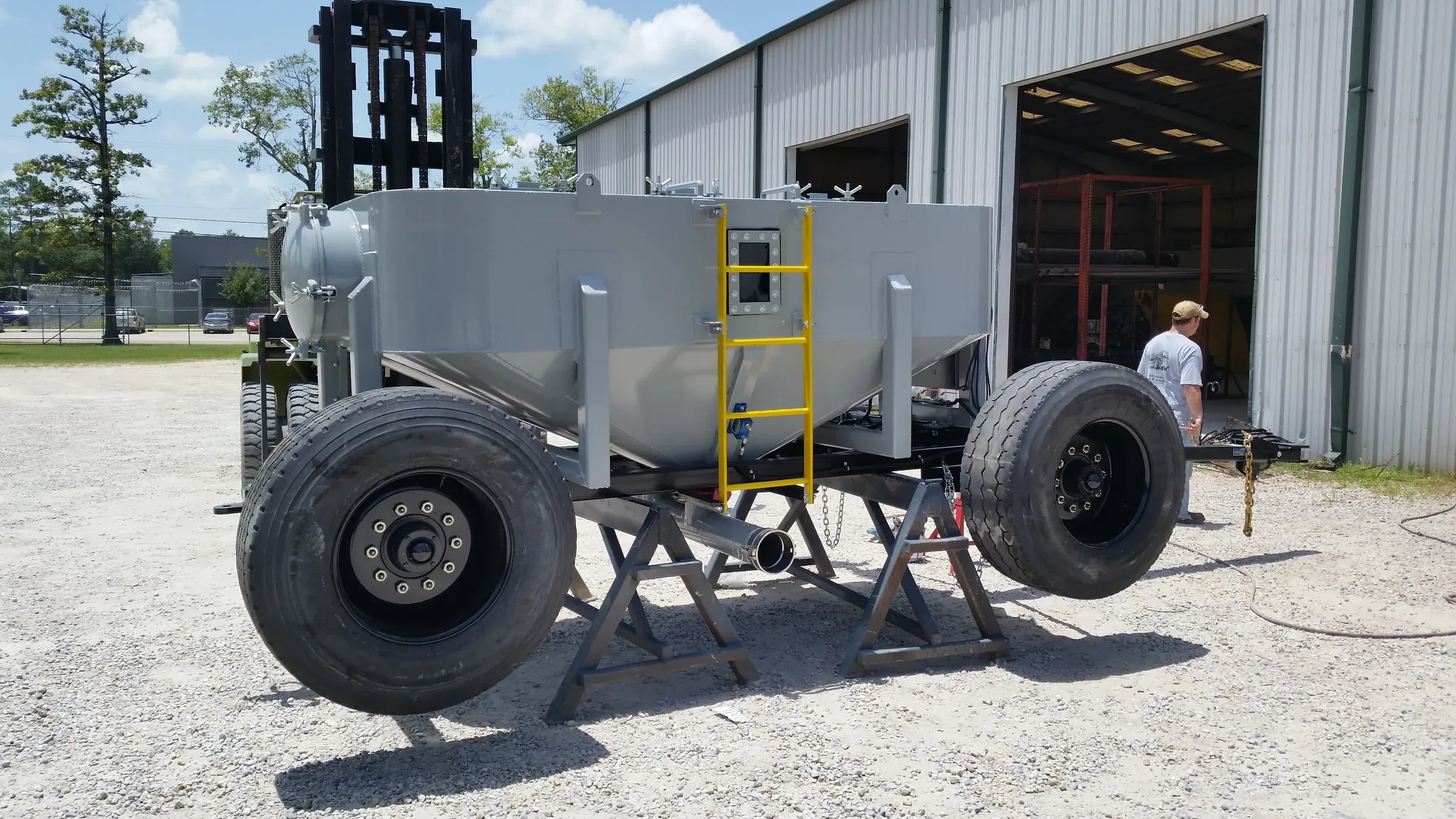 Completed grey tank trailer supported on stands in the gravel yard, technician walking past the open fabrication bay