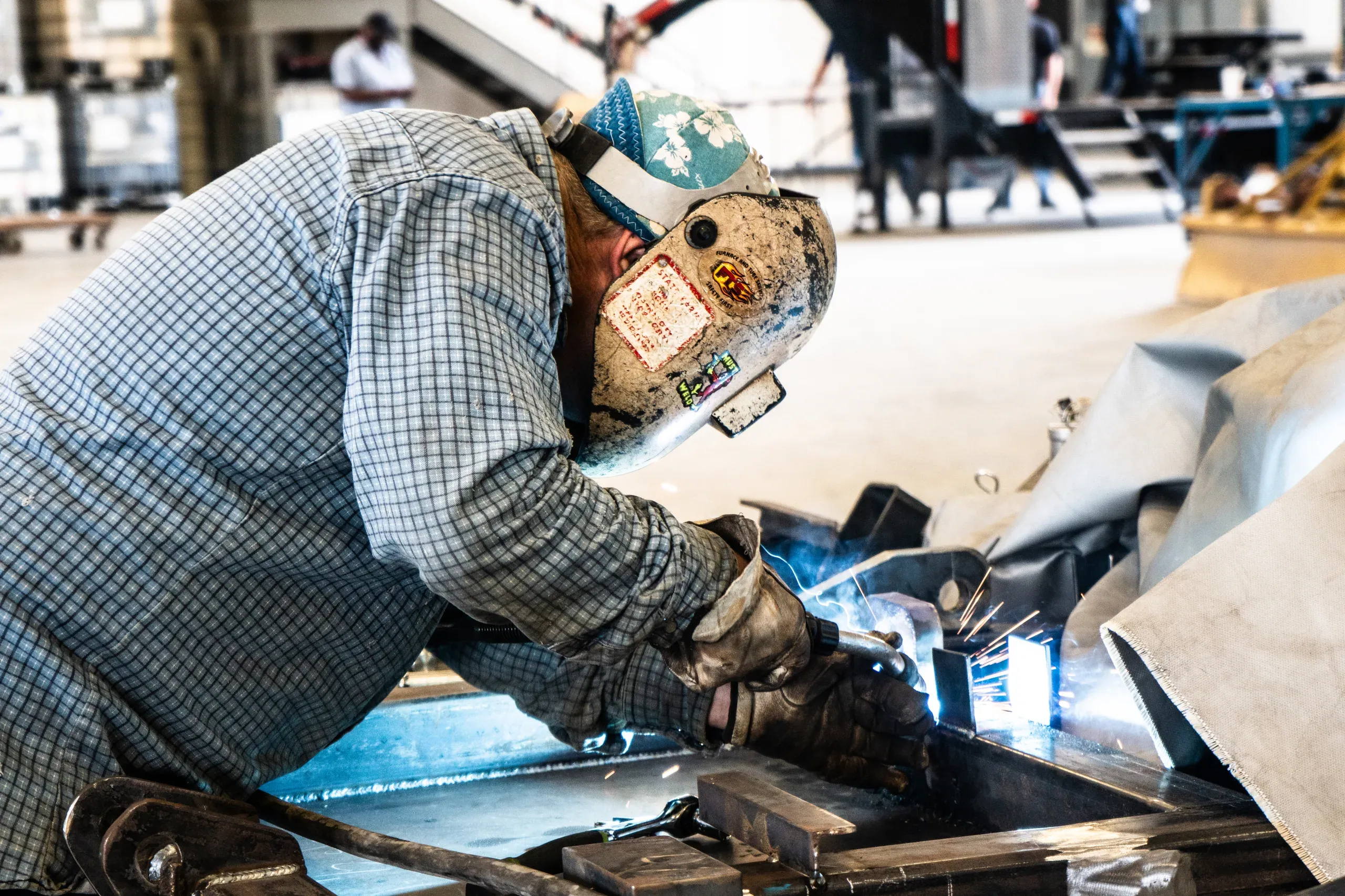 Welder in a sticker-covered hood running a bead along a steel weldment on the shop bench, arc glow reflecting off the plate while the shop works behind him