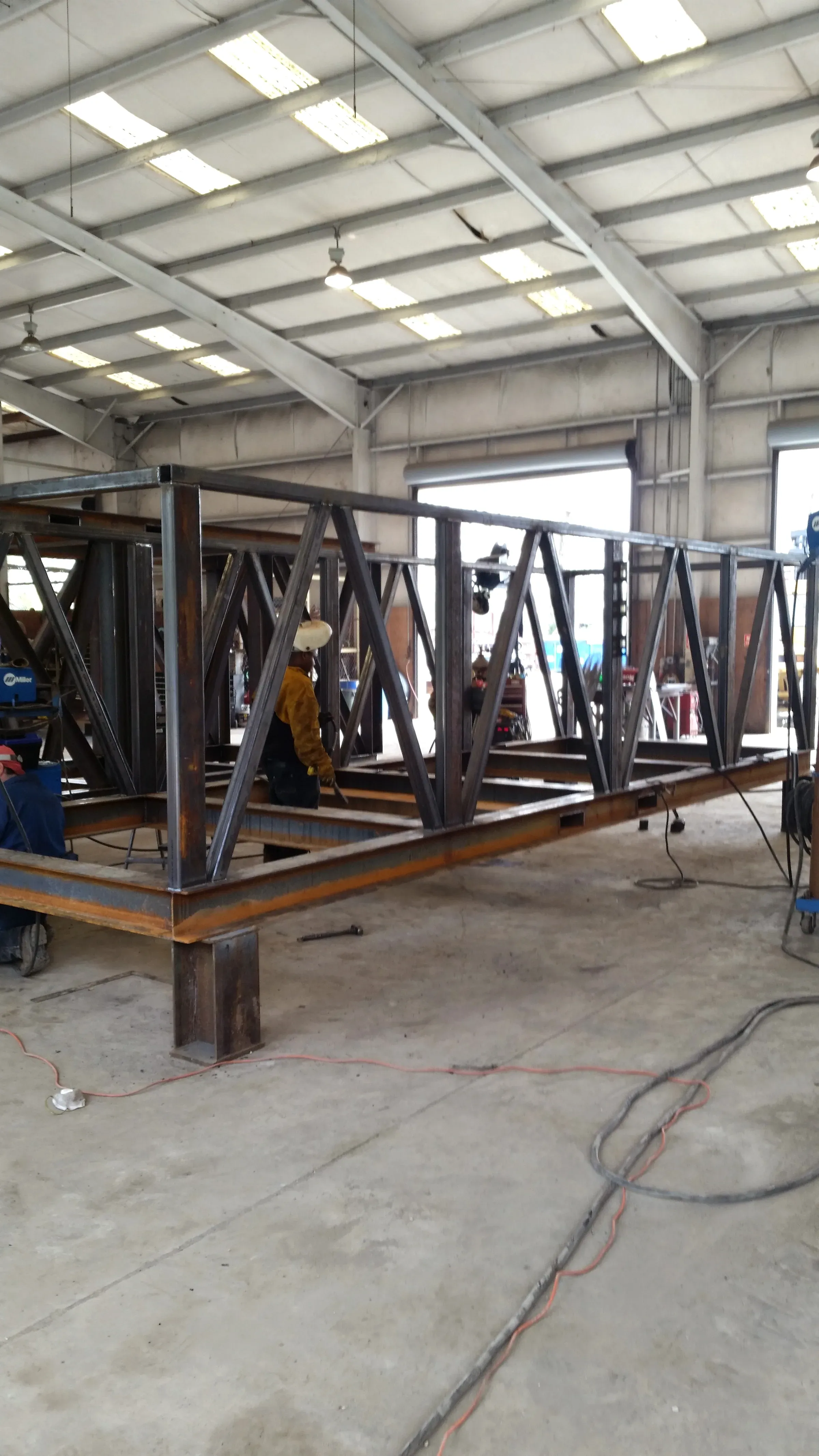 Welder in leathers and a hard hat working along a long cross-braced steel skid frame in the fabrication bay